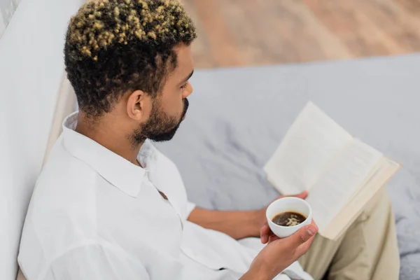Vue grand angle de jeune homme afro-américain avec des cheveux teints lecture roman et tenant tasse de café dans la chambre — Photo de stock