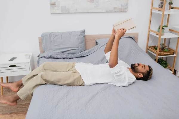 Giovane uomo africano americano con i capelli tinti tenendo libro sopra la testa e sdraiato sul letto — Foto stock