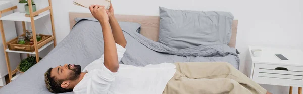 Joven afroamericano hombre con el pelo teñido sosteniendo libro sobre la cabeza y acostado en la cama, bandera - foto de stock