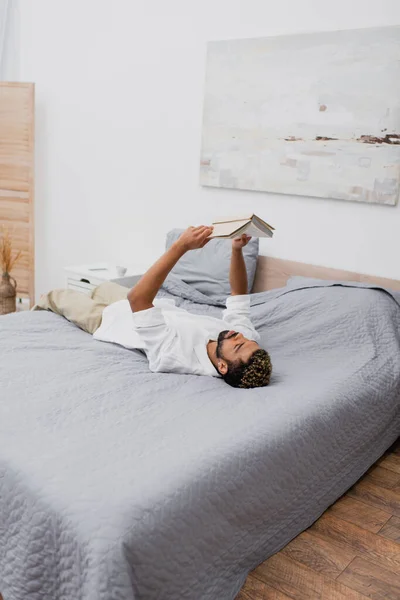 Jovem afro-americano com cabelo tingido segurando livro acima da cabeça e deitado na cama — Fotografia de Stock
