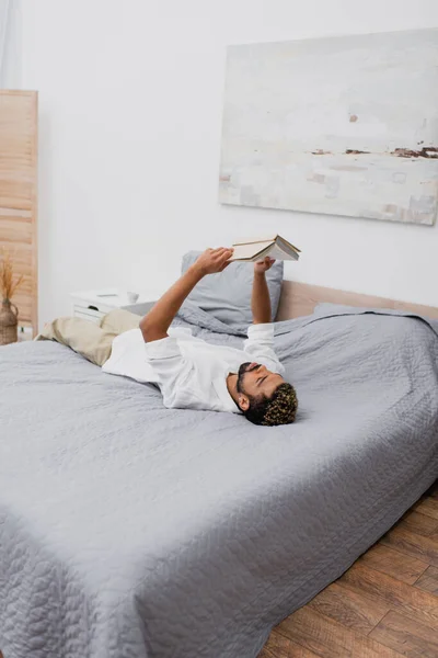 Giovane uomo africano americano con i capelli tinti tenendo libro sopra la testa e sdraiato sul letto — Foto stock