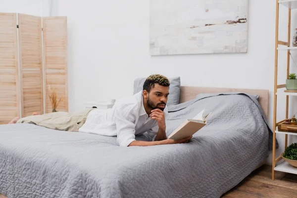 Junger afrikanisch-amerikanischer Mann mit gefärbten Haaren liest Buch während er auf dem Bett liegt — Stockfoto