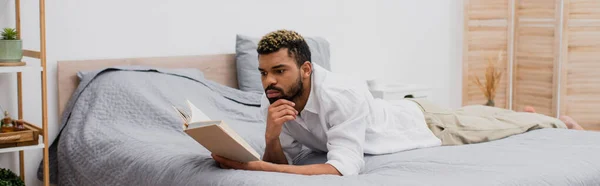 Africano americano hombre con teñido pelo lectura libro mientras está acostado en la cama, bandera - foto de stock