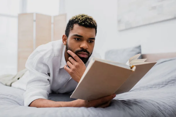 Joven afroamericano hombre con teñido pelo lectura novela mientras acostado en la cama - foto de stock