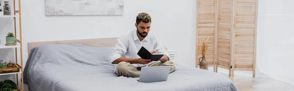 Jovem afro-americano homem segurando notebook e usando laptop enquanto trabalhava em casa no quarto, banner — Fotografia de Stock