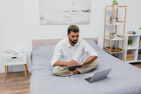 Jeune homme afro-américain tenant carnet et stylo tout en regardant ordinateur portable sur le lit — Photo de stock