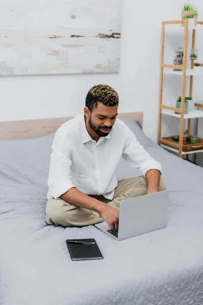 Young african american man smiling while using laptop on bed while working from home — Stock Photo