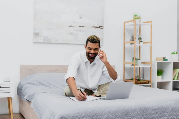 Hombre afroamericano feliz sonriendo mientras habla en el teléfono inteligente y escribir en portátil cerca de la computadora portátil en la cama - foto de stock