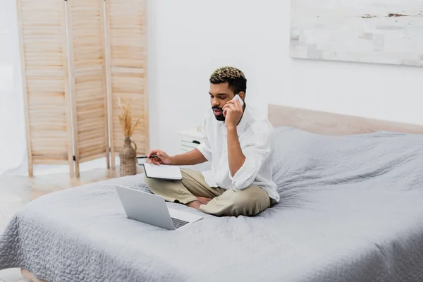 Hombre afroamericano hablando en el teléfono inteligente y la celebración de portátil con pluma cerca del ordenador portátil en la cama - foto de stock