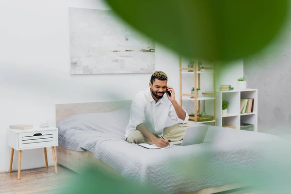 Heureux homme afro-américain parler sur smartphone tout en travaillant de la maison avec plante floue au premier plan — Photo de stock