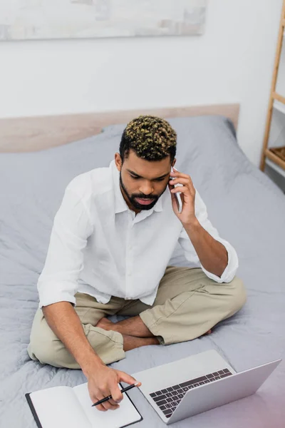 High angle view of young african american man sitting on bed while talking on smartphone and using laptop — Stock Photo