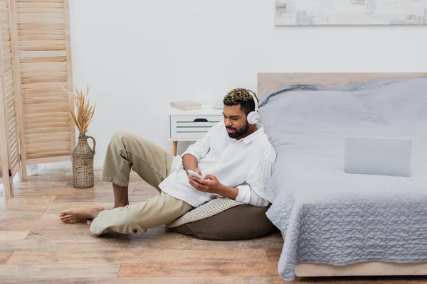 Молодой африканский американец в беспроводных наушниках, держащий смартфон, сидя рядом с ноутбуком на кровати — стоковое фото