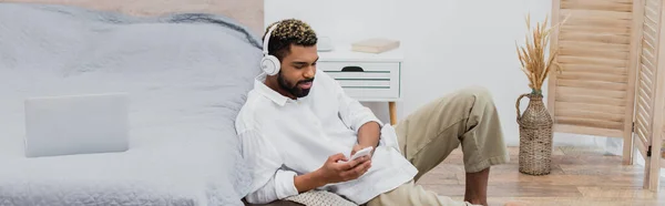 Young african american man in wireless headphones holding smartphone while sitting near laptop on bed, banner — Stock Photo