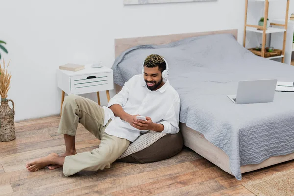 Felice uomo africano americano in cuffie senza fili utilizzando smartphone mentre seduto vicino al letto — Foto stock