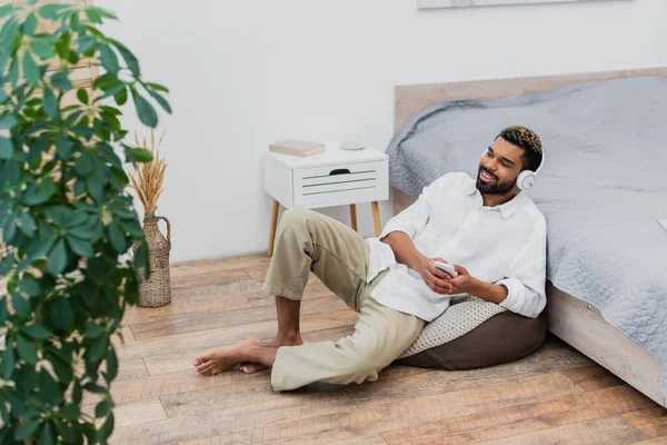 Heureux homme afro-américain dans les écouteurs sans fil écouter de la musique tout en tenant smartphone et assis près du lit — Photo de stock
