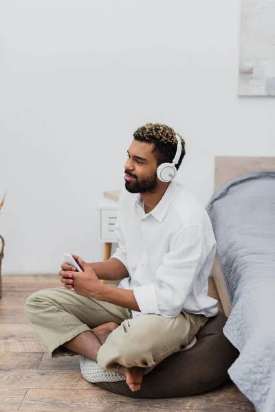 Cheerful african american man in wireless headphones listening music while holding smartphone and sitting near bed — Stock Photo