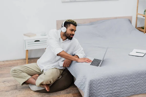 Joven afroamericano hombre en auriculares inalámbricos utilizando el ordenador portátil mientras sostiene el teléfono inteligente - foto de stock