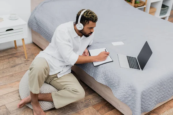 Jeune homme afro-américain dans les écouteurs sans fil écrit dans un cahier tout en regardant un ordinateur portable — Photo de stock