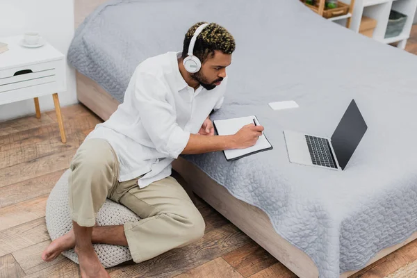 Giovane uomo afroamericano in cuffie senza fili che scrive nel notebook mentre guarda il computer portatile — Foto stock