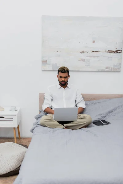 Jovem freelancer afro-americano usando laptop enquanto está sentado na cama — Fotografia de Stock