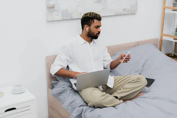Young african american man in wireless earphone using laptop and smartphone while sitting on bed — Stock Photo