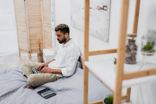 Giovane uomo africano americano in auricolare senza fili utilizzando il computer portatile mentre seduto sul letto a casa — Foto stock