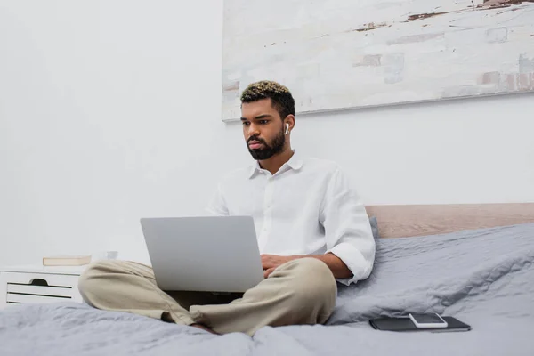 Молодий афроамериканський фрілансер в бездротових навушниках, використовуючи ноутбук, сидячи на ліжку вдома — стокове фото