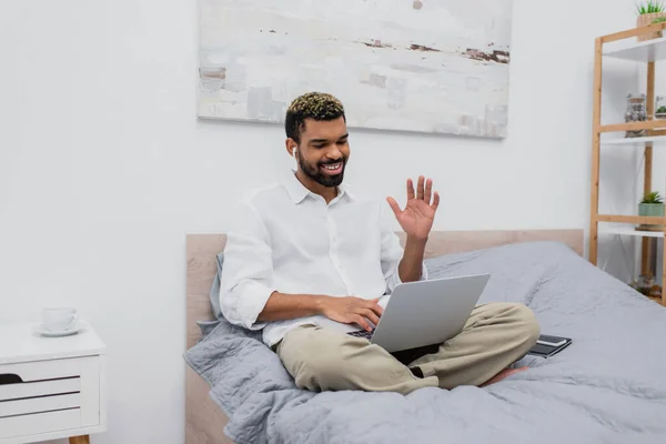 Hombre afroamericano feliz en auriculares inalámbricos agitando la mano durante la videollamada en el ordenador portátil mientras está sentado en la cama - foto de stock