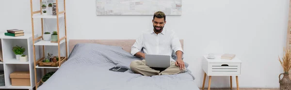 Homem americano africano alegre em fones de ouvido sem fio usando laptop enquanto sentado na cama, banner — Fotografia de Stock