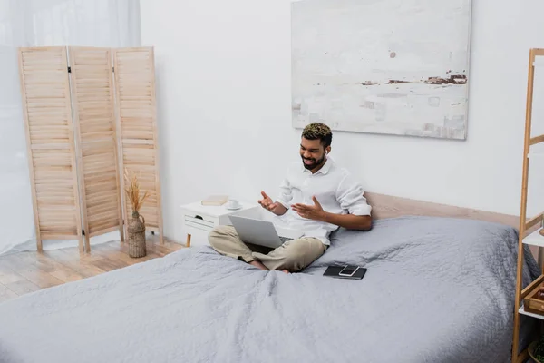 Homme afro-américain joyeux dans un écouteur sans fil ayant un appel vidéo sur un ordinateur portable tout en étant assis sur le lit — Photo de stock