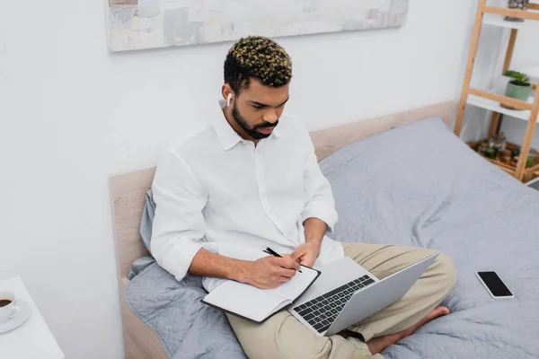 High angle view of african american man in wireless earphone writing on notebook and looking at laptop while sitting on bed — Stock Photo