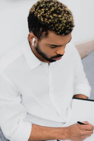 High angle view of young african american man in wireless earphone writing in notebook — Stock Photo