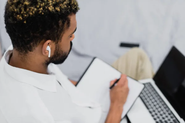 Young african american man in wireless earphone using laptop while writing in notebook — Stock Photo