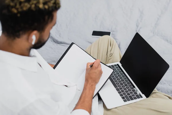 Visão de alto ângulo do homem americano africano desfocado em fone de ouvido sem fio usando laptop enquanto escreve em notebook — Fotografia de Stock