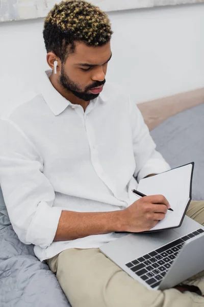 Joven afroamericano hombre en auriculares inalámbricos haciendo notas cerca de la computadora portátil en el dormitorio - foto de stock