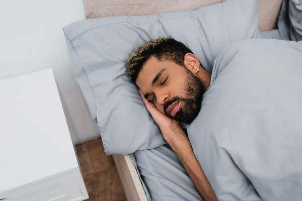 Visão de alto ângulo do homem americano africano barbudo com olhos fechados dormindo na cama perto da mesa de cabeceira — Fotografia de Stock