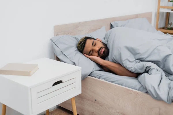 Homme afro-américain barbu avec les yeux fermés dormir dans le lit près de la table de chevet avec livre — Photo de stock