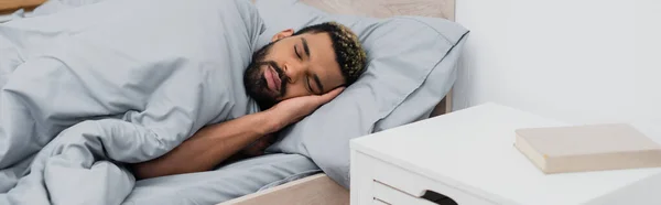 Homem americano africano barbudo com olhos fechados dormindo na cama perto da mesa de cabeceira com livro, banner — Fotografia de Stock