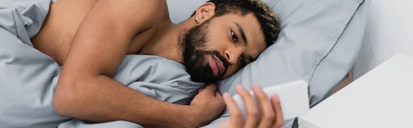 Barbudo hombre afroamericano usando teléfono inteligente mientras está acostado en la cama por la mañana, pancarta - foto de stock