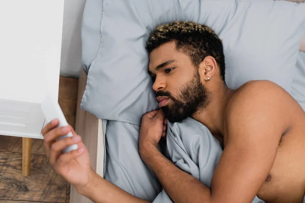 Vista superior do homem americano africano barbudo usando smartphone enquanto deitado na cama de manhã — Fotografia de Stock