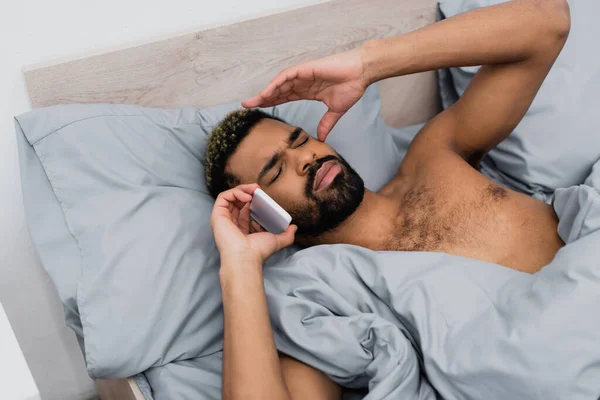 Visão de alto ângulo do homem americano africano despertado falando no smartphone na cama — Fotografia de Stock