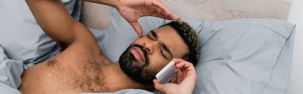 Visão de alto ângulo do homem americano africano despertado falando no smartphone na cama, banner — Fotografia de Stock