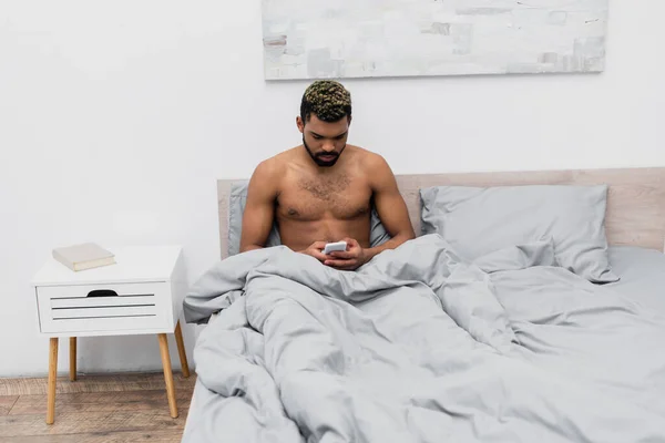 Hemdloser afrikanisch-amerikanischer Mann mit gefärbten Haaren chattet auf Smartphone im Schlafzimmer — Stockfoto