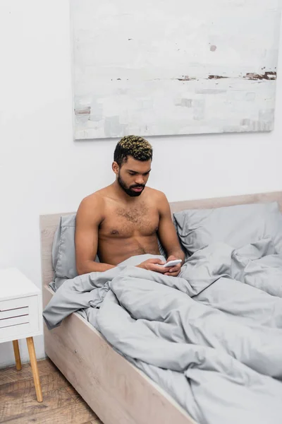 Uomo afroamericano senza maglietta con capelli tinti che chatta sul telefono cellulare a letto — Foto stock