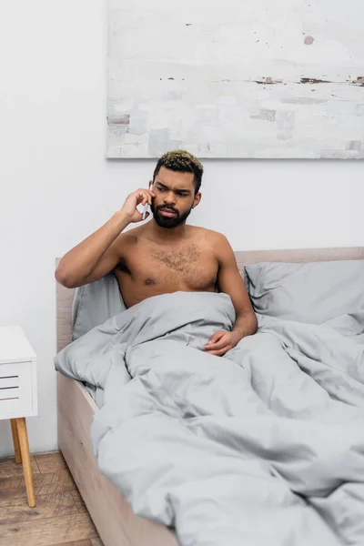 Hemdloser afrikanisch-amerikanischer Mann mit gefärbten Haaren, der im Bett mit dem Handy telefoniert — Stockfoto