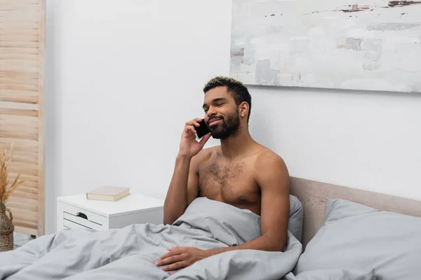 Homem americano africano sem camisa e feliz com cabelo tingido falando no telefone celular na cama — Fotografia de Stock