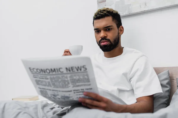 African american man with dyed hair holding cup of coffee while reading newspaper in bed — Stock Photo