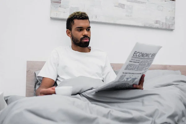 Barbuto uomo afroamericano con i capelli tinti in possesso di una tazza di caffè durante la lettura di un giornale a letto — Foto stock