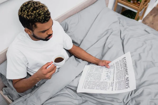 High angle view of african american man with dyed hair holding cup of coffee while reading newspaper in bed — Stock Photo