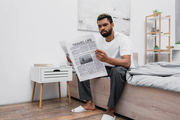 Hombre afroamericano con el pelo teñido sentado en la cama y leyendo el periódico de la vida de viaje - foto de stock